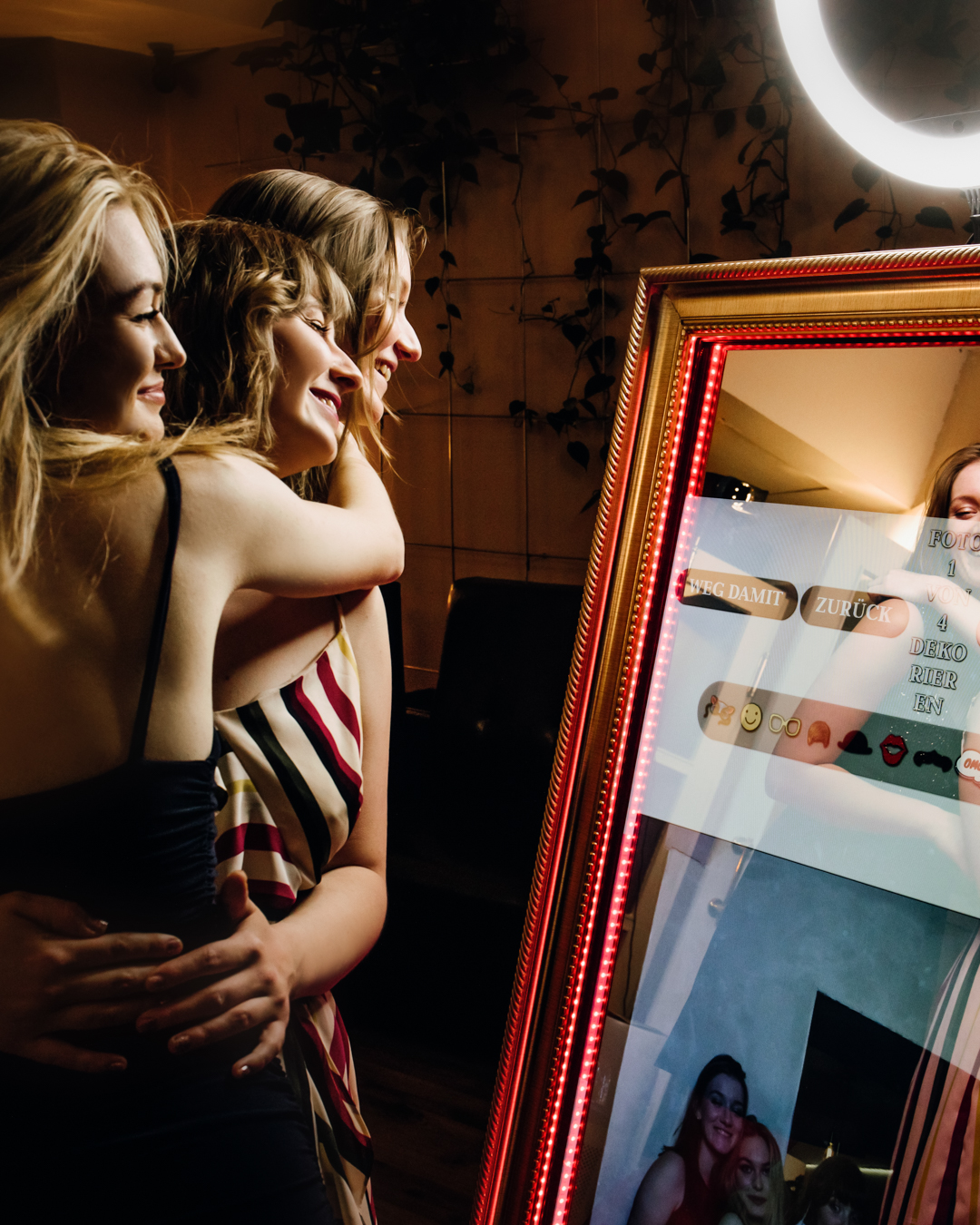 Drei fröhliche Frauen genießen Momente vor dem leuchtenden Fotospiegel (Fotobox), lachend und interagierend, in einem elegant dekorierten Raum.