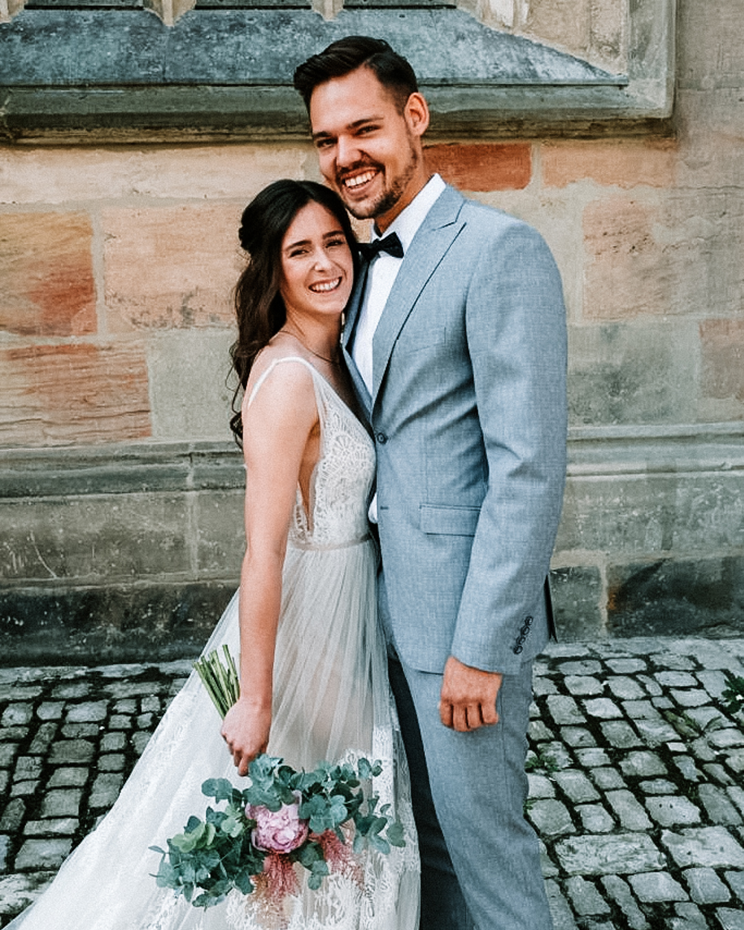 Strahlendes Brautpaar in Hochzeitskleidung lächelt vor historischer Steinmauer, die Braut hält einen Bouquet mit Eukalyptus und Pfingstrosen.