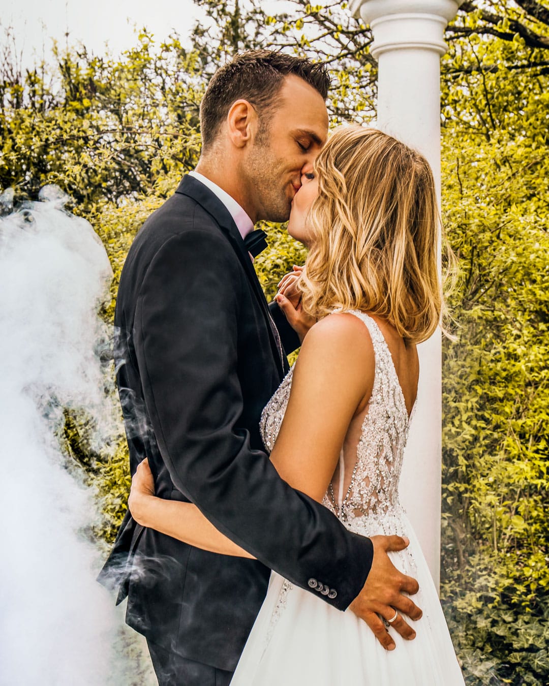 Strahlendes Brautpaar teilt einen liebevollen Kuss umgeben von nebeligem Ambiente bei einer Glüx-Hochzeitsfeier.