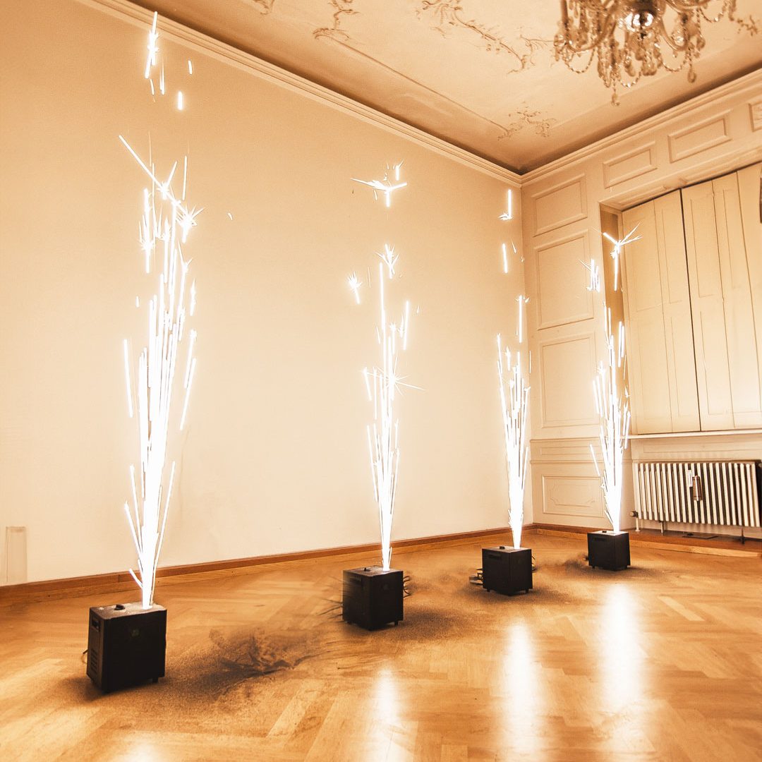 Funkenflug-Effekte von Glüx aus Funkensprüher-Geräten in einem eleganten Raum mit Holzboden, Stuckdecke und Kronleuchter, erzeugen kaltes Feuerwerk für eine Innenevent-Dekoration.