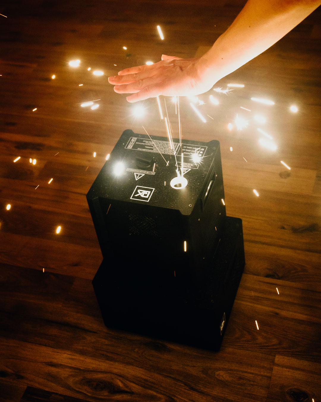 Ein Funkenflug-Gerät von Glüx auf einem Holzboden produziert kaltes Feuerwerk. Eine Hand nähert sich vorsichtig den aufsteigenden Funken, um die Ungefährlichkeit der Funken zu demonstrieren.