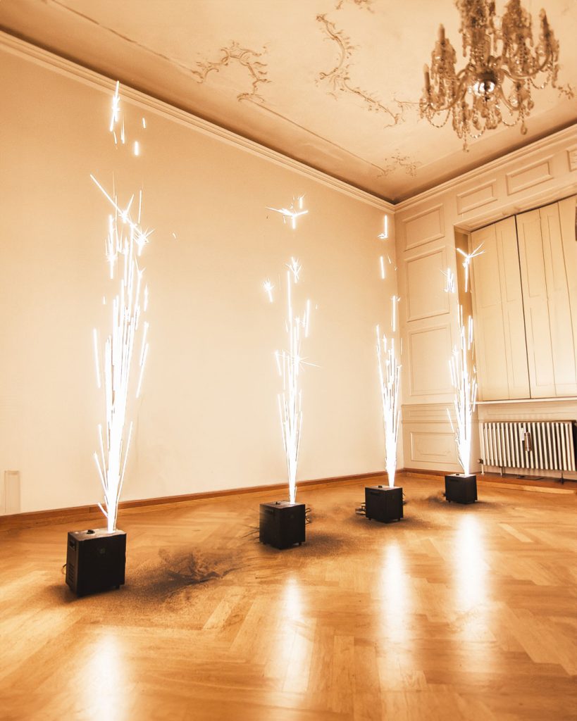 Funkenflug-Effekte von Glüx aus Funkensprüher-Geräten in einem eleganten Raum mit Holzboden, Stuckdecke und Kronleuchter, erzeugen kaltes Feuerwerk für eine Innenevent-Dekoration.