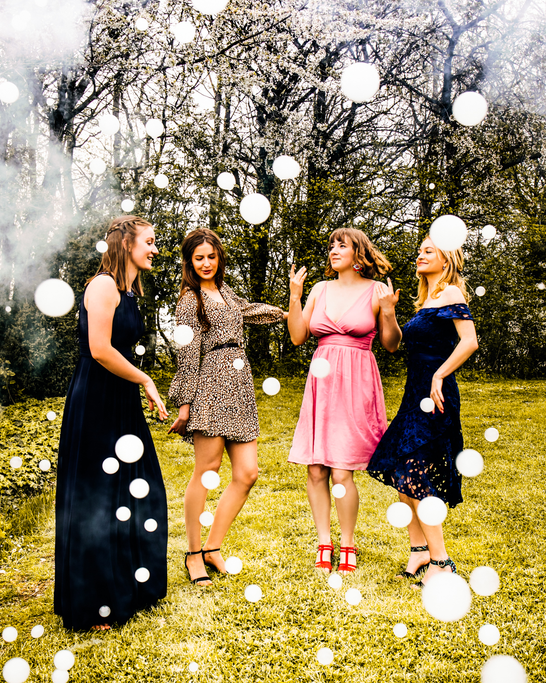 Vier Frauen in festlicher Kleidung haben Spaß in einem Garten, umgeben von einzigartigen weißen Seifenblasen (Nebelzauber) gefüllt mit Nebel. Die Bäume im Hintergrund sind voller Blüten, was eine fröhliche und feierliche Atmosphäre unterstreicht.